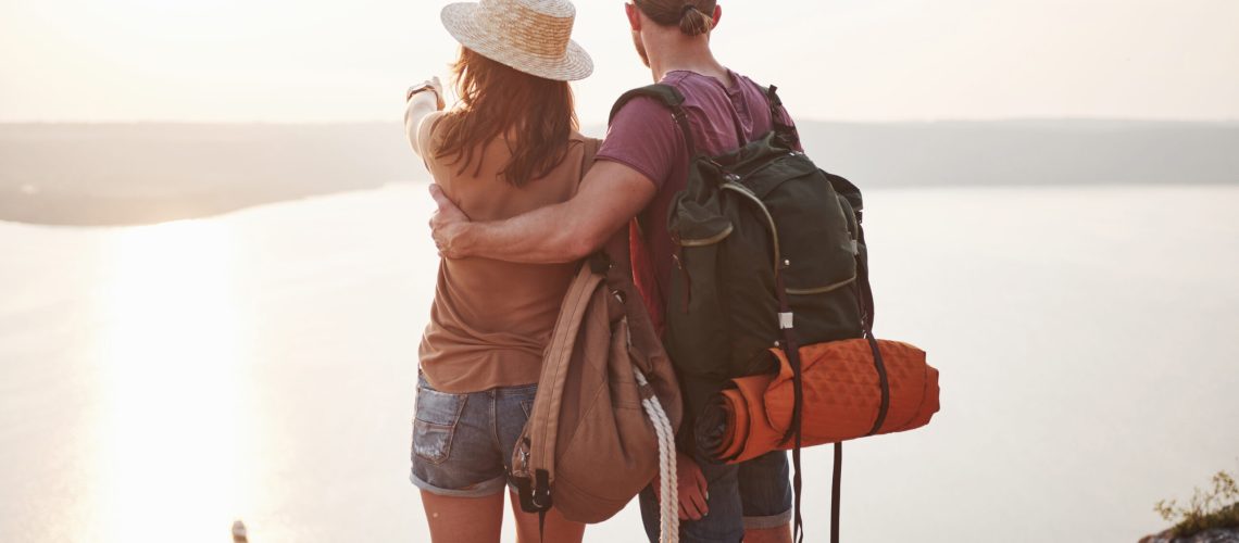 Two tourist male and woman with backpacks stand to the top of the mountain and enjoying sunrise. Travel lifestyle adventure vacations concept.